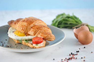 Arugula ile gevrek kruvasan, kızarmış yumurta, domates dilim. Çerçevede bir fincan kahve, bir demet arugula ve çatal bıçak takımı var. Açık arka plan.