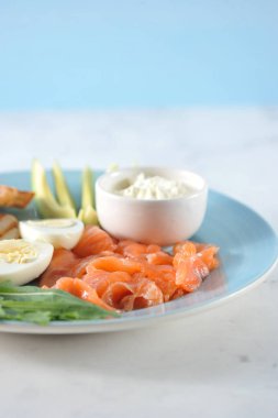 Kırmızı tuzlu balık, tost, haşlanmış yumurta, arugula, yumuşak krem peynir, mavi bir tabakta kireç ve avokado dilimleri. Brunch için ayarlayın. Yakınlarda çatal bıçak takımı var. Açık arka plan. Close. 