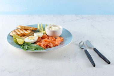 Kırmızı tuzlu balık, tost, haşlanmış yumurta, arugula, yumuşak krem peynir, mavi bir tabakta kireç ve avokado dilimleri. Brunch için ayarlayın. Yakınlarda çatal bıçak takımı var. Açık arka plan. Close. 