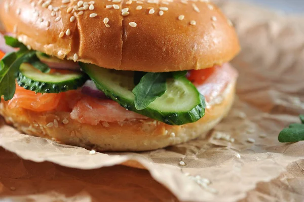 Beagle with a filling of salted salmon, arugula, cucumber, soft cheese. Beigle on kraft paper. On kraft paper sesame seeds. Light background. Close-up. Macro photography.