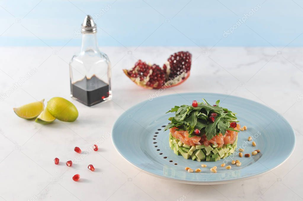 Tartar of salmon with arugula and avocado. Tar-tar on a blue plate is decorated with pine nuts. In the frame, slices of lime, pomegranate, balsamic sauce. Light background. Close-up.