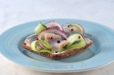 Mavi tabakta ringa balığı olan bir sandviç. Siyah ekmek krem peynir, genç ringa balığı, taze salatalık, soğan halkaları, kırmızı ve karabiber. Açık arka plan. Close. Makro fotoğrafçılık. 