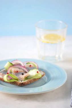 Mavi tabakta ringa balığı olan bir sandviç. Siyah ekmek krem peynir, genç ringa balığı, taze salatalık, soğan halkaları, kırmızı ve karabiber. Çerçevede, bir bardak su. Dikey çekim.