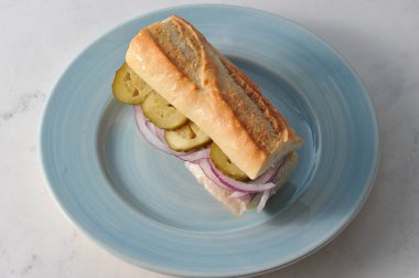 Genç ringa balığı ile geleneksel bir sandviç. Ringa balığı baget filetoları, salatalık turşusu dilimleri, soğan halkaları. Mavi tabakta sandviç. Açık arka plan. Close.