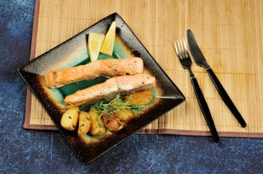 Somon bifteği ve tabakta kızarmış patates dilimleri. Tabakta, bezelye sürgünlerinden yeşillikler ve limon dilimleri. Plakanın yanında çatal bıçak takımı var. Tabağın altında bambudan yapılmış bir peçete var.. 