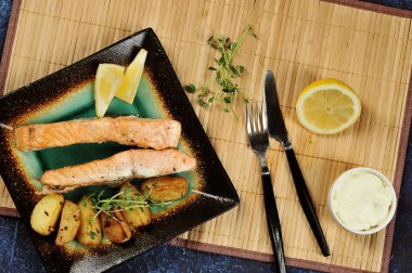 Somon bifteği ve kare tabakta kızarmış patates dilimleri. Bir sonraki çatal bıçak takımı, yarım limon, yumuşak peynirli bir fincan. Bambudan yapılmış plaka peçete altında bir arka plan oluşturur. Yukarıdan görüntüleyin. Close.