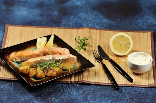 Somon bifteği ve kare tabakta kızarmış patates dilimleri. Bir sonraki çatal bıçak takımı, yarım limon, yumuşak peynirli bir fincan. Tabağın altında bambudan yapılmış bir peçete var. Karanlık arka plan. Close.