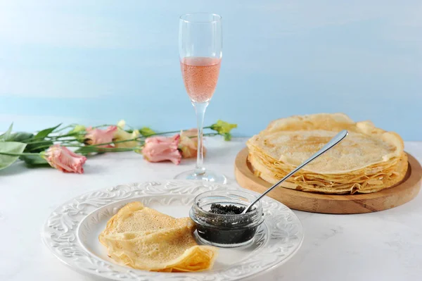 Thin pancakes on a wooden tray. Next to the plate with pancakes and black caviar. In the background, pink flowers and a glass of pink champagne. Vertical orientation of the frame. Light background.