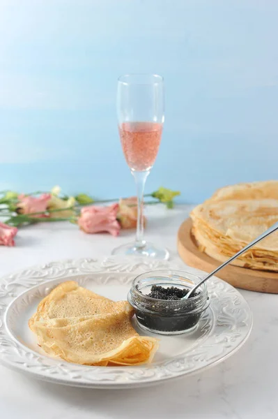 Thin pancakes on a wooden tray. Next to the plate with pancakes and black caviar. In the background, pink flowers and a glass of pink champagne. Vertical orientation of the frame. Light background.