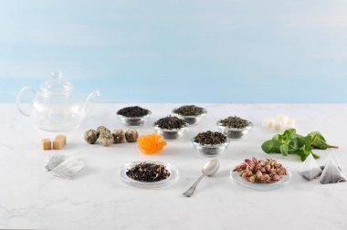 Çeşitli çay ve çaydanlık. Çerçevesiyah büyük yaprak çay, yeşil, yasemin ile yeşil, sütlü oolong, aromalı, topları çay. Nane yaprakları, kurutulmuş çiçek tomurcukları, şeker, çay poşetleri. Açık arka plan.