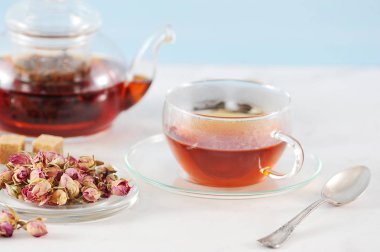 Siyah çaylı çaydanlık ve bir fincan çay. Cam eşyalar. Tabağın yanında kurutulmuş gül tomurcukları vardır, çay demlemek için kullanılır. Çerçevede, bir çay kaşığı. Açık arka plan. Close.