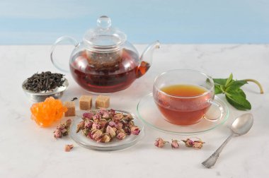 Siyah çaylı çaydanlık ve bir fincan çay. Cam eşyalar. Tabağın yanında kurutulmuş gül tomurcukları vardır, çay demlemek için kullanılır. Çerçeve, kaynak, nane, baston ve kristal şeker. Açık arka plan. Close.