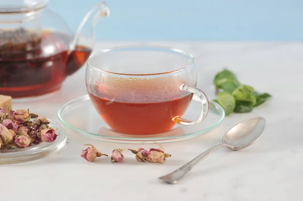 A teapot with black tea and a cup of tea. Glass ware. Next to the plate are dried rosebuds, used for brewing tea. In the frame, mint, a teaspoon. Light background. Close-up.