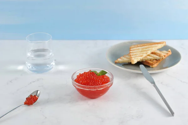 Red caviar and toast of white bread. Caviar in a glass cup, toast on a plate. Near the glass with water, a spoon with caviar. Light marble background. Close-up.