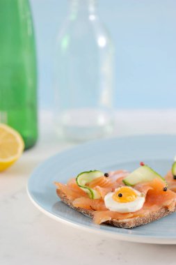 Somonlu sandviçler. Sandviç taze salatalık dilimleri, haşlanmış yumurta, soğan halkaları ile dekore edilmiştir. Arka planda su dolu bir bardak, bir şişe, bir limon. Açık arka plan. Dikey çekim.