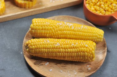 Yuvarlak ahşap bir tabakta iki mısır koçan. Mısır tuz büyük kristalleri serpilir. Arka planda, bir kase konserve mısır çekirdeği. Close. Gri arka plan.