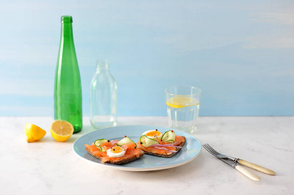 Sandwiches with salmon. The sandwich is decorated with fresh cucumber slices, hard-boiled egg, onion rings. Next to cutlery, a glass of water. In the background a bottle, a lemon. Light background.