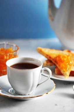 Bir fincan siyah çay ve bir tabak. Tabağın yanında tost ve portakal reçelli vazo var. Çerçevede, bir çaydanlık. Açık arka plan. Close. Makro fotoğrafçılık. Çerçevenin dikey yönü.