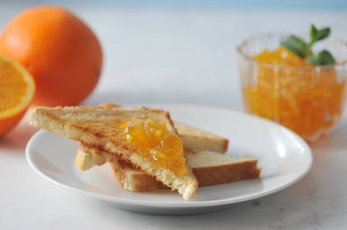 Portakal reçeli ve tost ile tabak. Çerçevede, bir portakal ve yarım portakal. Bir tost reçel ile bulaşmış. Tabağın yanında bir çay kaşığı var. Açık arka plan. Close. Makro fotoğrafçılık.