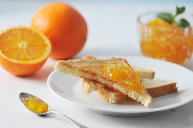 Portakal reçeli ve tost ile tabak. Çerçevede, bir portakal ve yarım portakal. Bir tost reçel ile bulaşmış. Tabağın yanında reçelli bir çay kaşığı var. Açık arka plan. Close. Makro fotoğrafçılık.