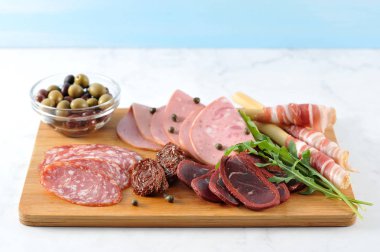 Tahta tepside, etli atıştırmalık. Haşlanmış dana sosisi, salchichen, kurutulmuş sığır-basturma, domuz pastırmalı grissini-breadsticks. Çerçevede, kurutulmuş domates, arugula, kapari, zeytin. Close.