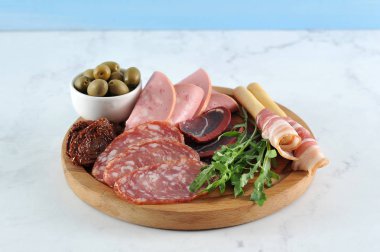 Yuvarlak tepside, etli atıştırmalık. Haşlanmış dana sosisi, salchichen, kurutulmuş sığır-basturma, domuz pastırmalı grissini-breadsticks. Çerçevede, kurutulmuş domates, arugula, kapari, zeytin. Close.