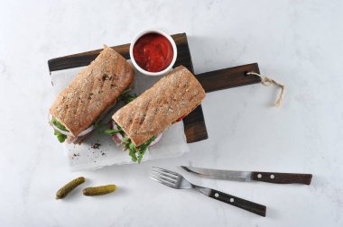 Tahta bir tahtada sandviç. Sandviç dolgu sığır eti, arugula, kırmızı soğan ve marine salatalık dilimleri oluşur. Çerçevede, salatalık, bir fincan ketçap, çatal bıçak takımı. Yukarıdan görüntüle.