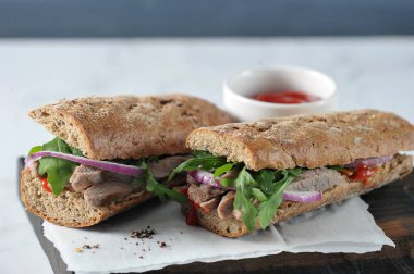 Tahta bir tahtada sandviç. Sandviç dolgu sığır eti, arugula, kırmızı soğan ve marine salatalık dilimleri oluşur. Çerçevede, bir fincan ketçap var. Close. Açık arka plan.