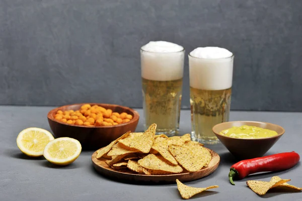 İki bardak hafif bira ve atıştırmalıklar. Tabaklarda, çıtır baharatlı kabukta yer fıstığı, mısır cipsi nachos, Meksika guacamole sosu. Plakaların yanında iki yarım kireç var. Gri arka plan.