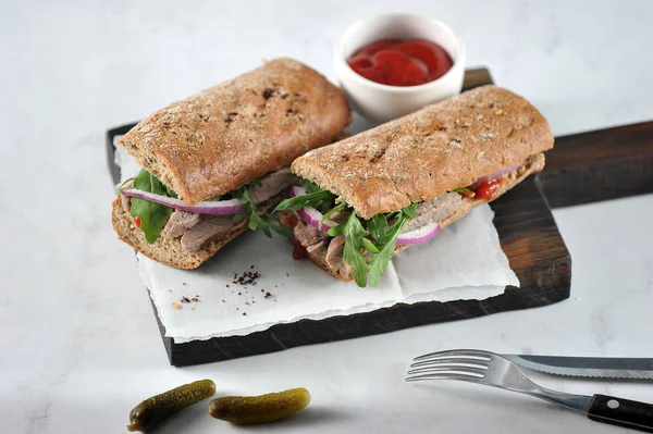 Sandwich on a wooden board. The filling of the sandwich consists of beef, arugula, red onion and marinated cucumber slices. In the frame, cucumbers, cup of ketchup, cutlery. Light background. Close-up