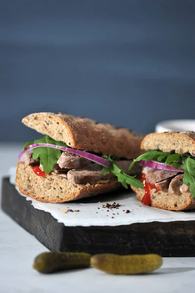 Sandwich on a wooden board. The filling of the sandwich consists of beef, arugula, red onion and marinated cucumber slices. Near the sandwich two cucumbers. Vertical shot. Negative space.