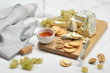 Hafif bir arka plan üzerinde peynir tabağı. Ahşap bir tabakta, küflü peynir, küllü camembert, brie, üzüm, kraker, bal ve ceviz. Çerçevede beyaz şarap ve bir tirbuşon ile bir bardak. Close.