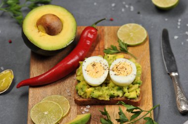 Avokado ve haşlanmış yumurta iki yarısı ile tost. Tost tahta bir tahtanın üzerinde yatıyor. Avokado, limon dilimleri, kırmızı biber, sarımsak, maydanoz çerçevesinde. Karanlık arka plan. Close. 