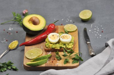 Avokado ve haşlanmış yumurta iki yarısı ile tost. Tost tahta bir tahtanın üzerinde yatıyor. Avokado, limon dilimleri, kırmızı biber, sarımsak, maydanoz çerçevesinde. Karanlık arka plan. Close. 