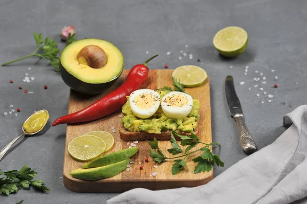 Avokado ve haşlanmış yumurta iki yarısı ile tost. Tost tahta bir tahtanın üzerinde yatıyor. Avokado, limon dilimleri, kırmızı biber, sarımsak, maydanoz çerçevesinde. Karanlık arka plan. Close. 