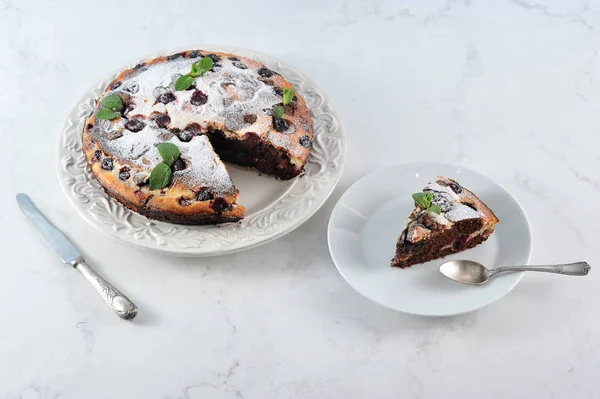 Curd-chocolate cake with cherries. The pie is decorated with powdered sugar and mint. On a separate plate, a piece of cake. Light marble background. Close-up.