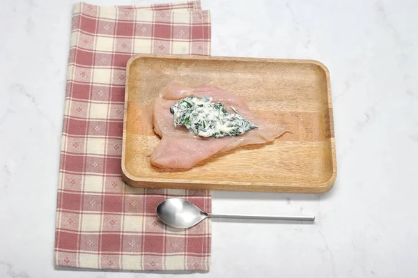 Stage preparation of chicken breast with spinach and cottage cheese filling. On a wooden plate a fillet with a mass of spinach and curd cheese.  In the frame there is a tablespoon. Light background.