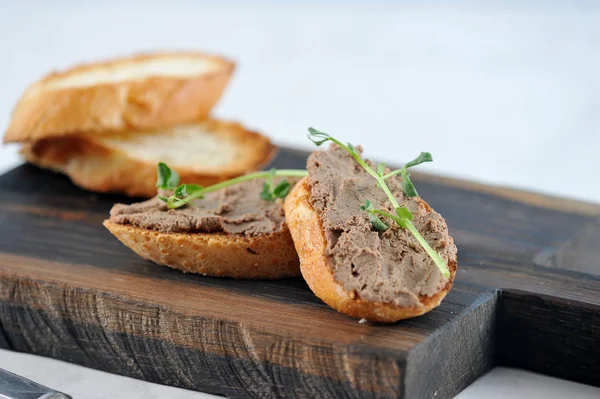 Kızarmış beyaz ekmek dilimleri üzerinde karaciğer pate. Sandviçler genç bezelye dalları ile dekore edilmiştir. Çerçevede ahşap bir tahta ve baget dilimleri bulunmaktadır. Açık arka plan. Close. Makro fotoğrafçılık.