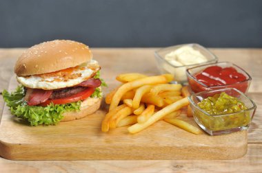 Açık ahşap bir yüzeyde hamburger. Burger pastırma, kızarmış yumurta ve kurutulmuş soğan içerir. Hamburgerin yanında patates kızartması ve soslar var. Close. 