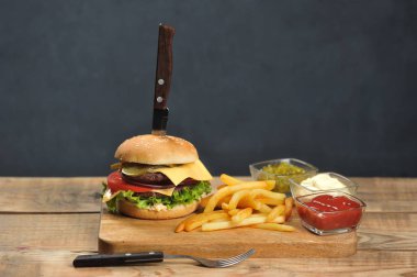 Hafif ahşap bir masada çift çizburger. Patates kızartması ve sos kaselerinin yanında. Hamburgerin içinde bıçak saplanmış. Close. Koyu arka plan. 