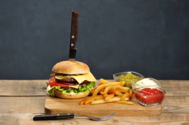 Hafif ahşap bir masada çift çizburger. Patates kızartması ve sos kaselerinin yanında. Hamburgerin içinde bıçak saplanmış. Close. Koyu arka plan. 