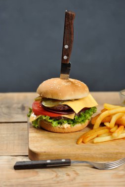 Hafif ahşap bir masada çift çizburger. Patates kızartması ve sos kaselerinin yanında. Hamburgerin içinde bıçak saplanmış. Close. Karanlık arka plan. Dikey çerçeve yönlendirmesi.