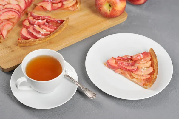 Tabakta bir parça elmalı turta. Bir fincan çayın yanında. Arka planda elmalı turta ve elma vardır. Gri arka plan. Close.