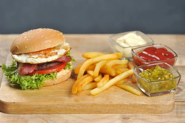 Açık ahşap bir yüzeyde hamburger. Burger pastırma, kızarmış yumurta ve kurutulmuş soğan içerir. Hamburgerin yanında patates kızartması ve soslar var. Close. 