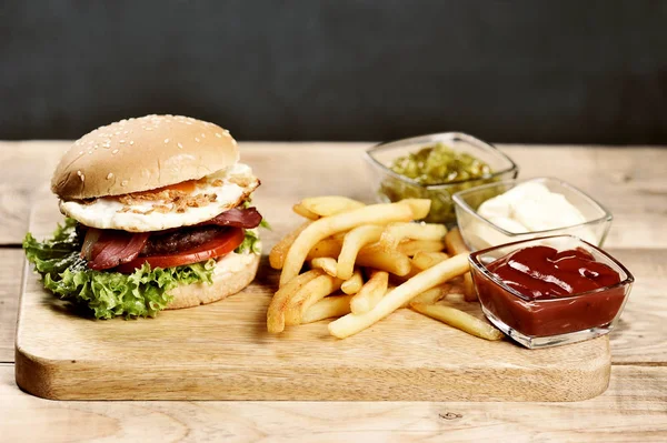 Hafif ahşap tahtada hamburger. Burger pastırma, kızarmış yumurta ve kurutulmuş soğan içerir. Burgerin yanında patates kızartması ve soslu bardaklar var. Close.
