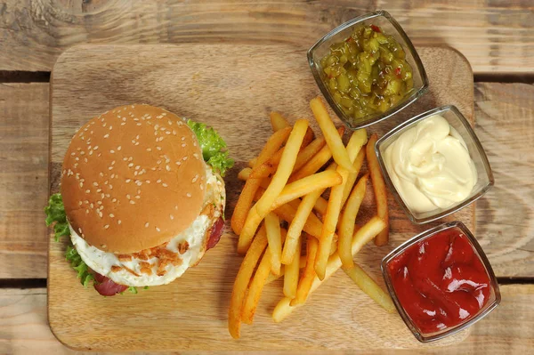 Hamburger on a light wooden surface. The burger includes bacon, fried egg and dried onions. Next to the burger are french fries and sauces. Close-up. View from above.