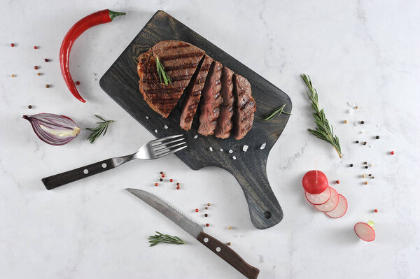 On a dark wooden board a juicy steak cooked on the grill. In the frame cutlery, fresh radish, rosemary, chili pepper. View from above. Light background. Close-up.