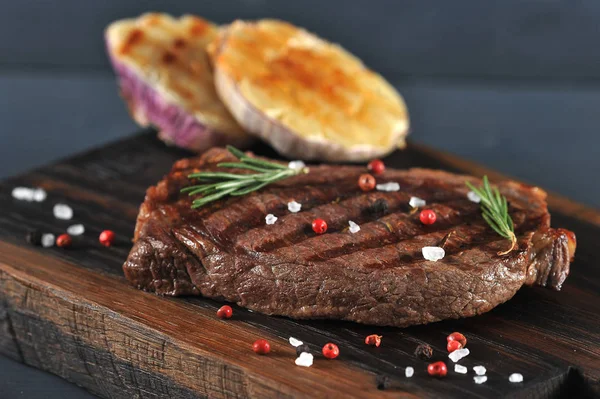 On a wooden board, beef steak cooked on the grill. Next to the steak are two halves of garlic, cooked on the grill. In the frame a cup with ketchup.Close-up. Macro photography. Dark background.