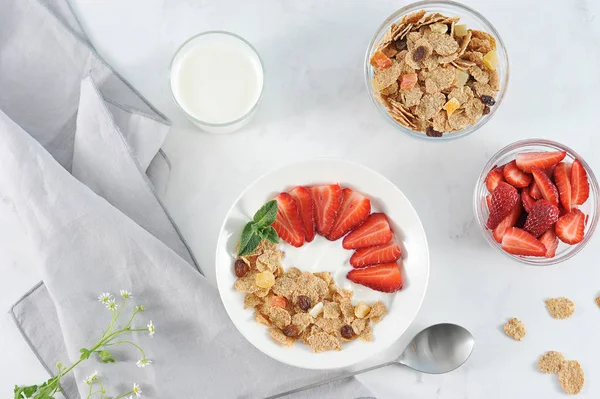 Plate with corn flakes with the addition of fresh strawberry yogurt. Next to the glass with yogurt, cups with cereal and strawberries. Light background. Close-up. View from above.