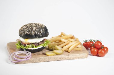 Hafif ahşap tahtada siyah burger. Dolgu köfte, kızarmış yumurta, sos, sebze oluşur. Hamburger kızartmasının yanında. Arka planda bir kiraz domates dalı. Açık arka plan. Close.
